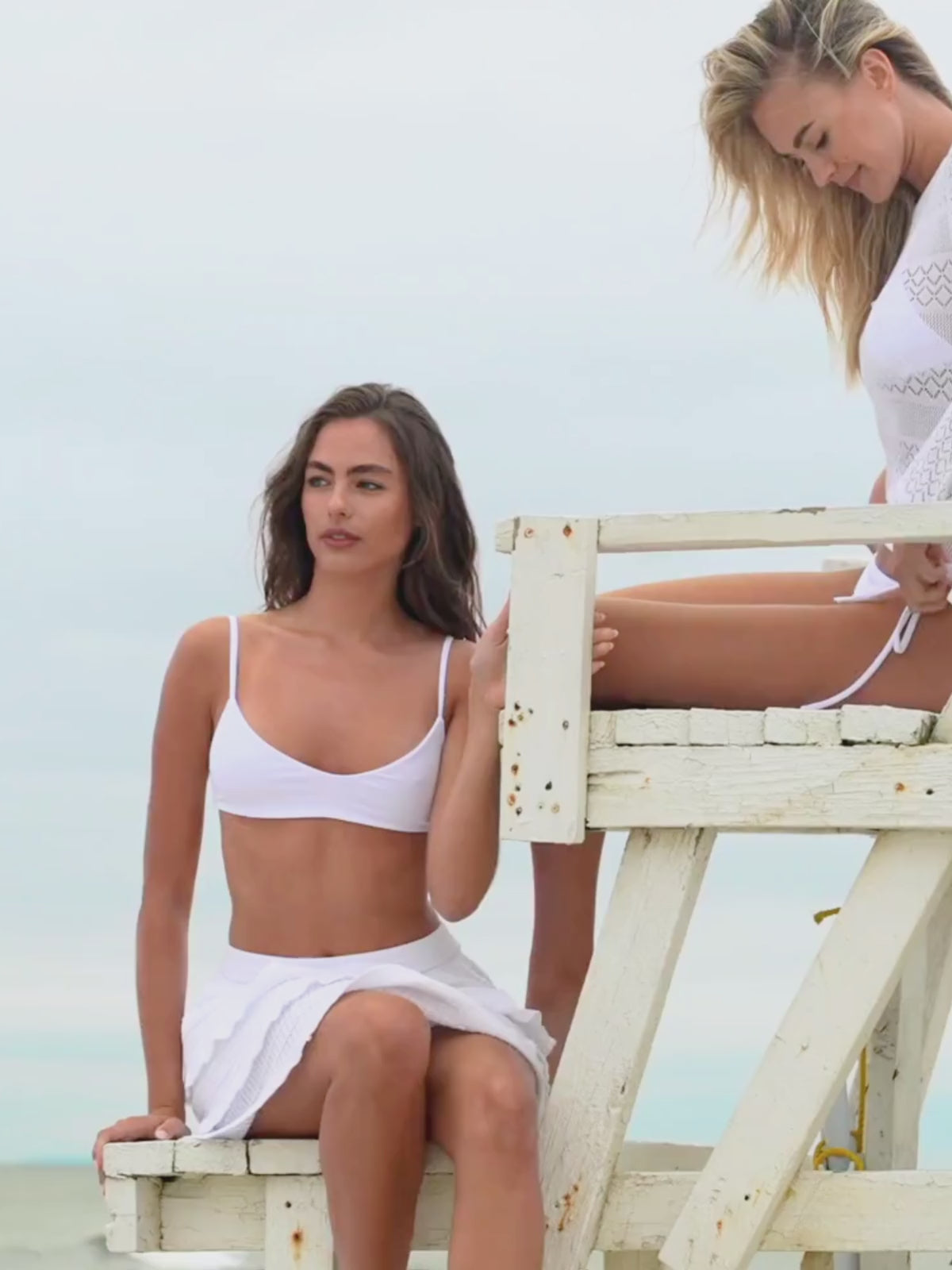 Two Woman one wearing a white long-sleeve pointelle sweater top with a tie bikini  another wearing bralette and pointelle skirt on a beach background